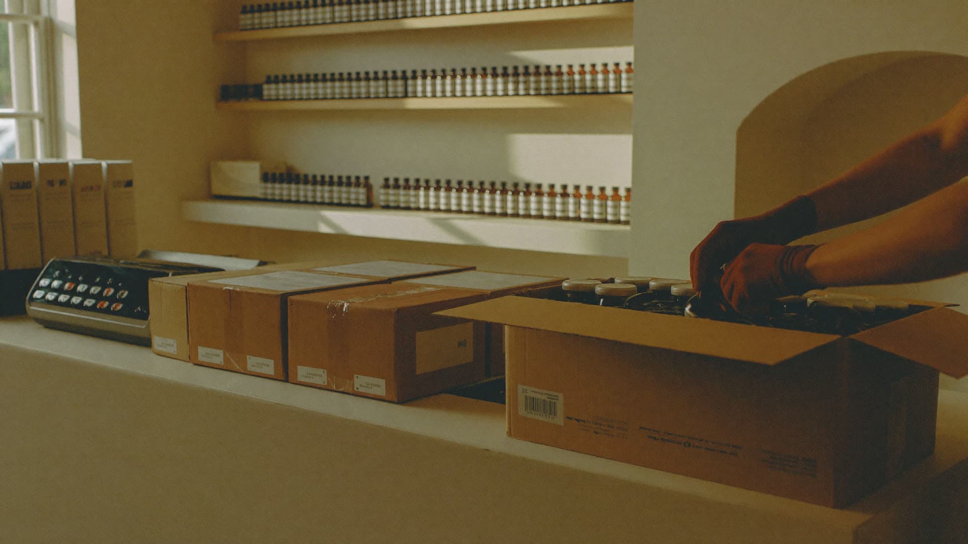 Gloved hands packing amber bottles into a kraft shipping box at a warm pharmacy bench, shelves of bottles behind